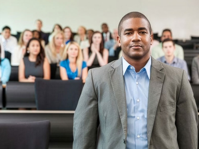 Person standing in a classroom with seated students in the background during a lecture.